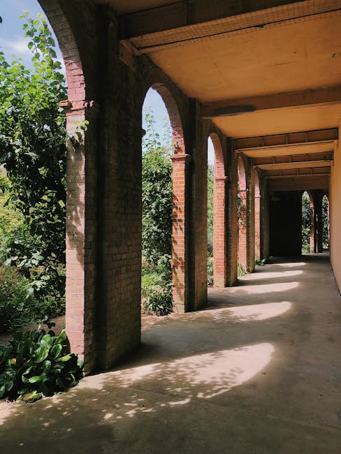 A covered outdoor walkway featuring a series of brick arches supported by sturdy brick columns, casting dappled shadows on the concrete floor. Lush green foliage and plants are visible along the left